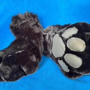 May include: A pair of black furry paw gloves with gray paw pads. The gloves are made of a soft, plush material and are designed to resemble animal paws. The gloves are lying on a blue surface.