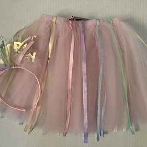 May include: A pink tulle skirt with colorful ribbons and a matching headband. The headband has the words "Happy Birthday" in gold. The skirt is designed for a child's birthday celebration.