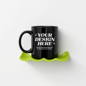 May include: A black coffee mug with the text "YOUR DESIGN HERE" in white, resting on a lime green, leaf-shaped coaster. The mug has a black handle and the text "instant digital mockup file to showcase your design."