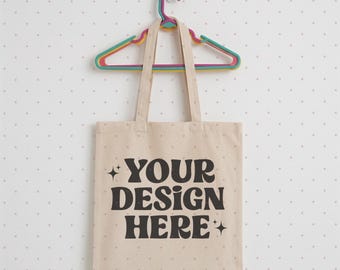 May include: A beige canvas tote bag with two shoulder straps hanging on a multi-colored plastic hanger. The bag features the black text "YOUR DESIGN HERE" printed in a bold, rounded font, flanked by two small four-pointed star icons on the left and right sides.