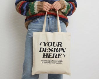 Puede incluir: Una bolsa de tela color crema con el texto impreso en negro "YOUR DESIGN HERE" en una fuente serif negrita, acentuada por iconos de estrellas de cuatro puntas, con un texto más pequeño que dice "Instant digital mockup file to showcase your design."