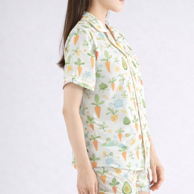 May include: A white two-piece pyjama set with a repeating pattern of orange carrots, green leaves, blue watering cans, and yellow flowers. The short-sleeved top has a collared neckline and a front pocket. The shorts have an avocado graphic.