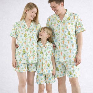 May include: Coordinated light blue pyjama sets with a garden-themed pattern. The sets include short-sleeved button-up shirts and shorts, featuring carrots, avocados, and watering cans. The pyjamas are worn by three people.