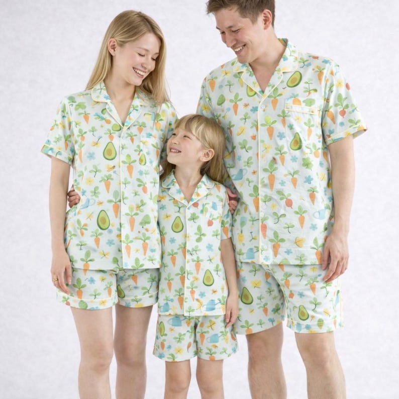 May include: Coordinated light blue pyjama sets with a garden-themed pattern. The sets include short-sleeved button-up shirts and shorts, featuring carrots, avocados, and watering cans. The pyjamas are worn by three people.
