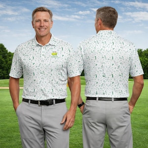 Op de afbeelding: Witte poloshirt met een groen golfpatroon en het Masters-logo. Het shirt wordt gedragen met een grijze broek en een zwarte riem. De afbeelding toont de voor- en achterkant van het shirt.