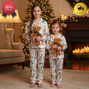 May include: Two girls wearing matching white pajamas with a festive print, holding brown teddy bears. The pajamas have red trim and a pattern of holiday-themed illustrations. A Christmas tree and fireplace are in the background.