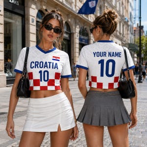 May include: Two women wearing white Croatia soccer jerseys with blue trim, the word "CROATIA" and the number "10" in blue. The jerseys have a red and white checkered pattern at the bottom. One woman wears a white skirt, the other a gray pleated skirt.