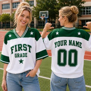 May include: A white and dark green V-neck jersey with short sleeves and striped trim. The front features the text "FIRST GRADE" in dark green letters with a star. The back displays "YOUR NAME" and the number "00" in dark green with stars.
