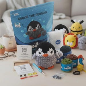 Könnte beinhalten: Ein Häkelset mit einem grau-schwarzen Pinguin mit orangefarbenem Schnabel, mit der Aufschrift "GOOKIE THE PENGUIN". Das Set enthält Garn, eine Häkelnadel und weiteres Zubehör. Weitere gehäkelte Tierfiguren sind ebenfalls zu sehen.