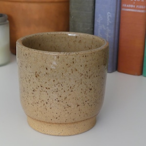 May include: A speckled beige ceramic planter with a cylindrical shape and a slightly raised base. The planter has a smooth, glossy finish and is suitable for indoor or outdoor use. The background includes a candle in a jar and a stack of books.