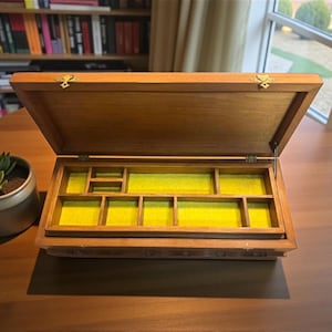 May include: Open wooden jewelry box with a hinged lid and brass-colored hardware. The interior is lined with yellow felt and divided into several compartments. A small potted succulent sits to the left of the box on a wooden table.