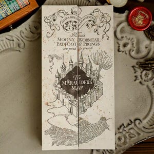 May include: A folded paper map with the text "The Marauder's Map" and the names "Moony, Wormtail, Padfoot & Prongs." The map is tan with black ink and features a castle illustration. The map is on a decorative white tray.