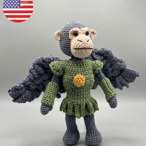 May include: A handmade crochet monkey doll with gray wings and a green outfit. The doll has a gray head and limbs, with a tan face and ears. A yellow circle is on the front of the green outfit. The doll is a unique, handcrafted item.