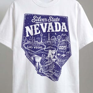Pode incluir: Uma t-shirt branca de manga curta com uma impressão gráfica azul escura do estado do Nevada. O design inclui o texto Silver State NEVADA, o sinal Welcome to Fabulous Las Vegas, uma slot machine, dados, um alienígena e uma paisagem desértica.