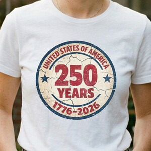 以下が含まれることがあります： アメリカ合衆国250周年を記念した円形デザインの白いTシャツ。「United States of America」、「250 Years」、および「1776-2026」の文字が赤と青でデザインされています。