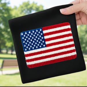 Könnte beinhalten: Ein schwarzes quadratisches Textil mit einem gestickten amerikanischen Flaggenmuster. Die Flagge zeigt blaue Sterne und rot-weiße Streifen. Das Textil wird von einer Hand vor einem verschwommenen grünen Hintergrund gehalten.
