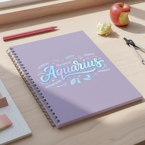 May include: A spiral-bound notebook with a lavender cover featuring the word "Aquarius" in a whimsical, multi-colored script. The design includes additional words like "Visionary" and "Creative". A pink pen and a partially eaten apple are also visible.