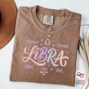 May include: A brown Comfort Colors t-shirt with a Libra design. The design includes the word "Libra" in pastel colors, along with the words "Harmony", "Balance", "Grace", and "Fair". A silver necklace with a pendant is on the shirt.