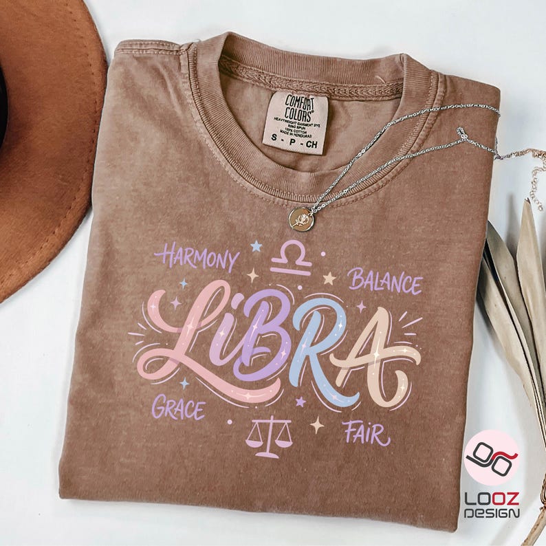 May include: A brown Comfort Colors t-shirt with a Libra design. The design includes the word "Libra" in pastel colors, along with the words "Harmony", "Balance", "Grace", and "Fair". A silver necklace with a pendant is on the shirt.