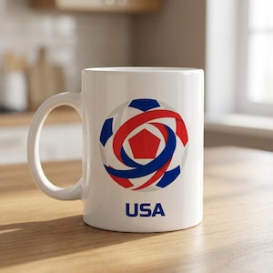 May include: White ceramic mug featuring a soccer ball design in red, blue, and white, with the word "USA" printed below. The mug has a curved handle and is set on a wooden surface.