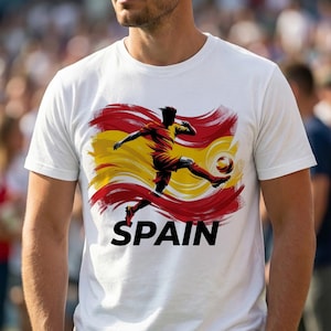 Puede incluir: Camiseta blanca con un jugador de f&uacute;tbol pateando un bal&oacute;n, sobre un fondo de pinceladas rojas, amarillas y naranjas. La palabra "SPAIN" est&aacute; impresa en negrita negra debajo. Prenda de vestir informal.