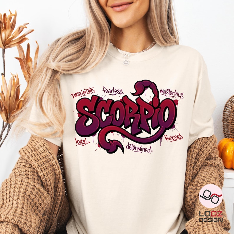 May include: Cream-colored t-shirt featuring a stylized Scorpio graphic in shades of purple and red. The word "Scorpio" is the focal point, surrounded by words like "passionate" and "mysterious". The design has a graffiti-style aesthetic.