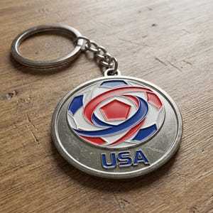 May include: A silver-colored keychain with a round pendant featuring a soccer ball design in red, white, and blue. The word "USA" is displayed in blue letters at the bottom of the pendant. The keychain is on a wooden surface.