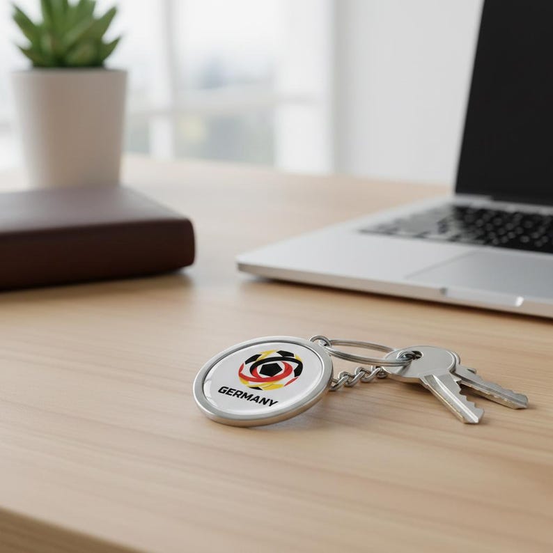 May include: A silver keychain with a round pendant featuring the German flag colors and a soccer ball design. The word "Germany" is printed below the graphic. Two keys are attached to the chain, resting on a light-colored wooden surface.
