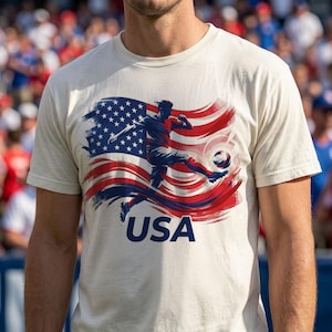 Puede incluir: Una camiseta color crema con un gr&aacute;fico de un jugador de f&uacute;tbol pateando un bal&oacute;n, sobre un fondo de la bandera estadounidense en rojo, blanco y azul. La palabra "USA" est&aacute; impresa en azul marino debajo. La camiseta es de manga corta.