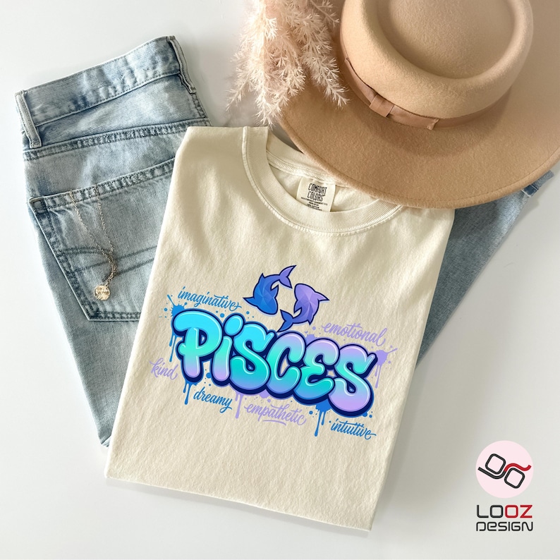 May include: Cream-colored t-shirt with a Pisces design in blue and purple. The design includes two fish and the word "Pisces" in a graffiti-style font, along with words like "imaginative" and "emotional". The shirt is paired with light-wash jeans and a tan hat.