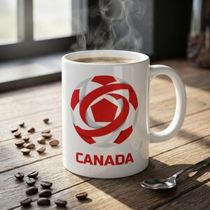 May include: A white ceramic coffee mug with a red and gray graphic design and the word "CANADA" in red. The mug is filled with coffee, and steam rises from the top. Coffee beans and a spoon are on the wooden surface.