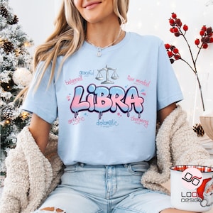 May include: Light blue t-shirt featuring the word "Libra" in a bubble letter style, with pink and blue dripping paint effect. The shirt also includes the words "balanced," "graceful," "fair minded," "artistic," "diplomatic," and "charming."