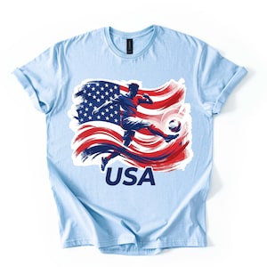 May include: Light blue t-shirt featuring a graphic of a soccer player kicking a ball, set against a stylized American flag in red, white, and blue. The word "USA" is printed in white below the graphic.