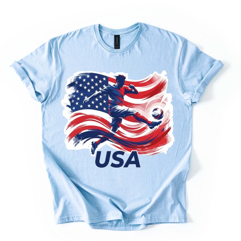 May include: Light blue t-shirt featuring a graphic of a soccer player kicking a ball, set against a stylized American flag in red, white, and blue. The word "USA" is printed in white below the graphic.
