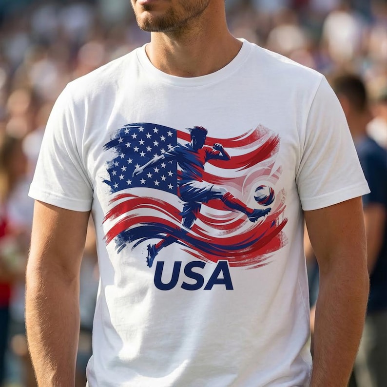 Puede incluir: Camiseta blanca con un jugador de f&uacute;tbol pateando un bal&oacute;n, sobre un dise&ntilde;o estilizado de la bandera estadounidense en rojo, blanco y azul. La palabra "USA" est&aacute; impresa en azul debajo.