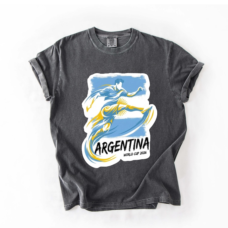 Może przedstawiać: Ciemnoszara koszulka z grafiką piłkarza w kolorze niebieskim i ż&oacute;łtym, z napisem "ARGENTINA" i "WORLD CUP 2026" w kolorze białym. Koszulka ma podwinięte rękawy i okrągły dekolt.