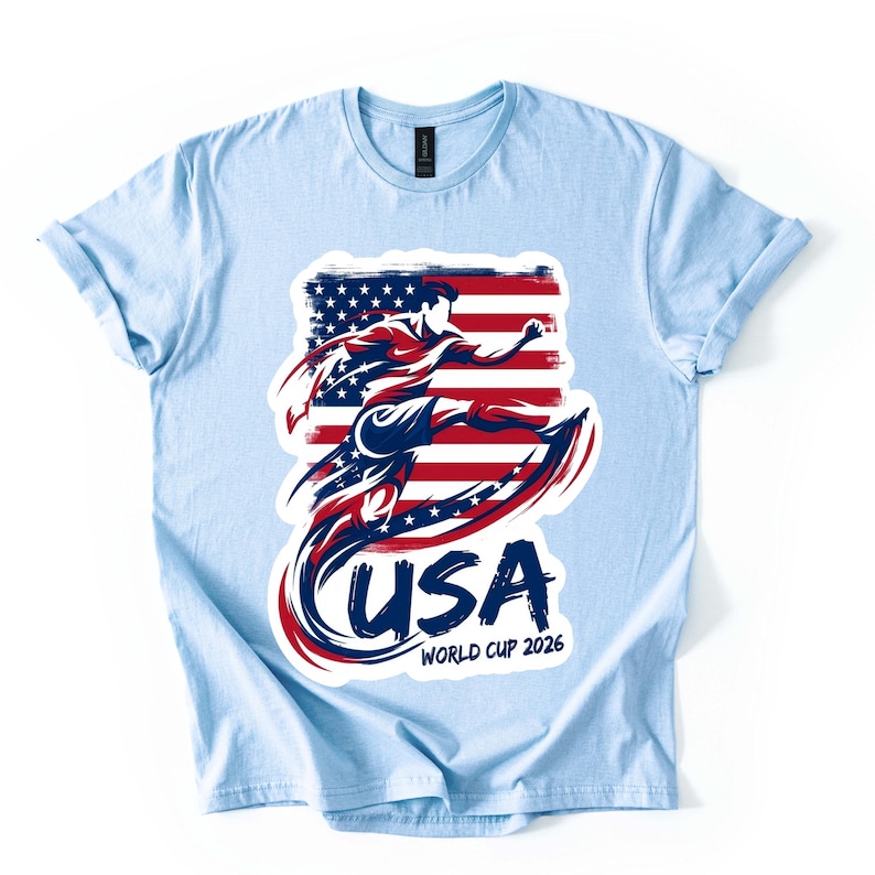 Może przedstawiać: Jasnoniebieska koszulka z grafiką piłkarza kopiącego piłkę, na tle flagi USA. Słowo "USA" jest wyraźnie wyświetlane, wraz z "WORLD CUP 2026". Koszulka ma podwinięte rękawy.