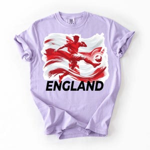 Puede incluir: Camiseta de color lila claro con un gr&aacute;fico de un futbolista pateando un bal&oacute;n, sobre un dise&ntilde;o de bandera roja y blanca. La palabra "ENGLAND" est&aacute; impresa en letras negras en negrita debajo del gr&aacute;fico. La camiseta tiene las mangas enrolladas.