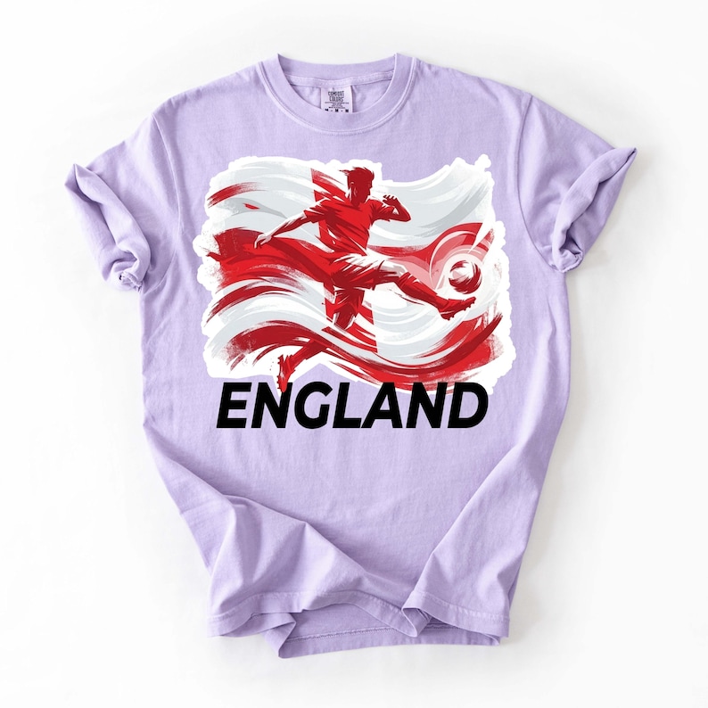 Puede incluir: Camiseta de color lila claro con un gr&aacute;fico de un futbolista pateando un bal&oacute;n, sobre un dise&ntilde;o de bandera roja y blanca. La palabra "ENGLAND" est&aacute; impresa en letras negras en negrita debajo del gr&aacute;fico. La camiseta tiene las mangas enrolladas.