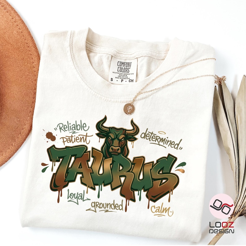 May include: Cream-colored t-shirt featuring a green and brown Taurus graphic with a bull illustration. The shirt includes the words "Reliable, Patient, Determined, Loyal, Grounded, Calm." A gold necklace and a brown hat are also visible.
