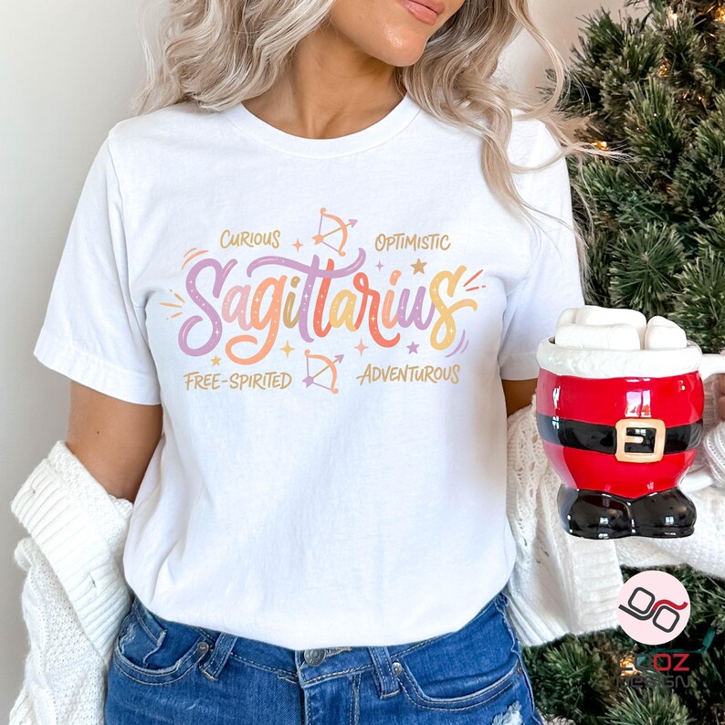 May include: White t-shirt with a colorful Sagittarius design. The design includes the word "Sagittarius" in a gradient of orange, pink, and yellow, along with the words "Curious", "Optimistic", "Free-Spirited", and "Adventurous".