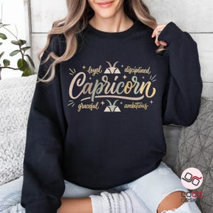 May include: A black sweatshirt featuring the word "Capricorn" in a stylish, multi-colored font. The design includes the words "loyal, disciplined, graceful, ambitious" with star and mountain graphics. A casual, comfortable top.