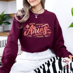 May include: Burgundy Aries sweatshirt with a colorful graphic design. The design includes the word "Aries" in a stylized font, along with the words "Passion, Confidence, Fire, Strength, Bold". The sweatshirt is made of a soft material.