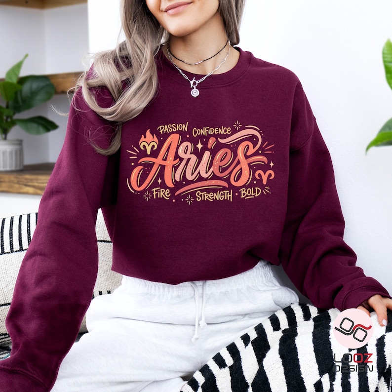 May include: Burgundy Aries sweatshirt with a colorful graphic design. The design includes the word "Aries" in a stylized font, along with the words "Passion, Confidence, Fire, Strength, Bold". The sweatshirt is made of a soft material.