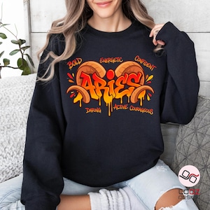 May include: Black sweatshirt featuring a colorful Aries design. The design includes the word "Aries" in orange and yellow graffiti style, with the words "Bold," "Energetic," "Confident," "Daring," "Active," and "Courageous" surrounding it.
