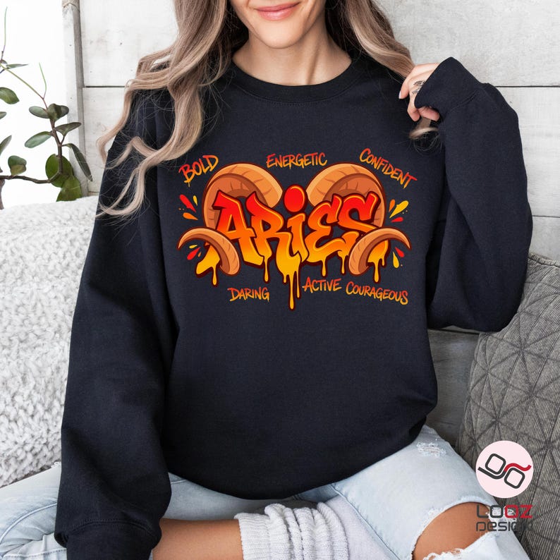 May include: Black sweatshirt featuring a colorful Aries design. The design includes the word "Aries" in orange and yellow graffiti style, with the words "Bold," "Energetic," "Confident," "Daring," "Active," and "Courageous" surrounding it.