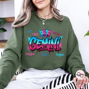 May include: An olive green sweatshirt with a colorful Gemini graphic. The design features the word "Gemini" in a graffiti style, with blue and pink accents, and the words "Witty", "Curious", "Expressive", "Clever", "Adaptable", and "Sociable".