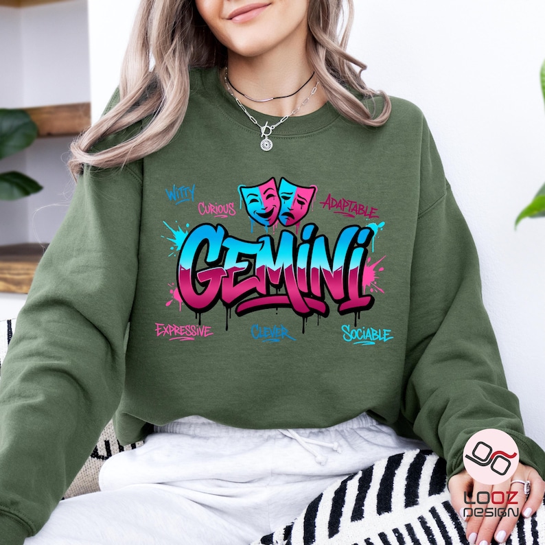 May include: An olive green sweatshirt with a colorful Gemini graphic. The design features the word "Gemini" in a graffiti style, with blue and pink accents, and the words "Witty", "Curious", "Expressive", "Clever", "Adaptable", and "Sociable".