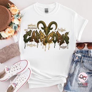 May include: White t-shirt featuring a Capricorn design with a goat illustration and the word "Capricorn" in a graffiti style. The design includes words like "ambitious", "disciplined", "wise", "practical", "focused", and "resilient".