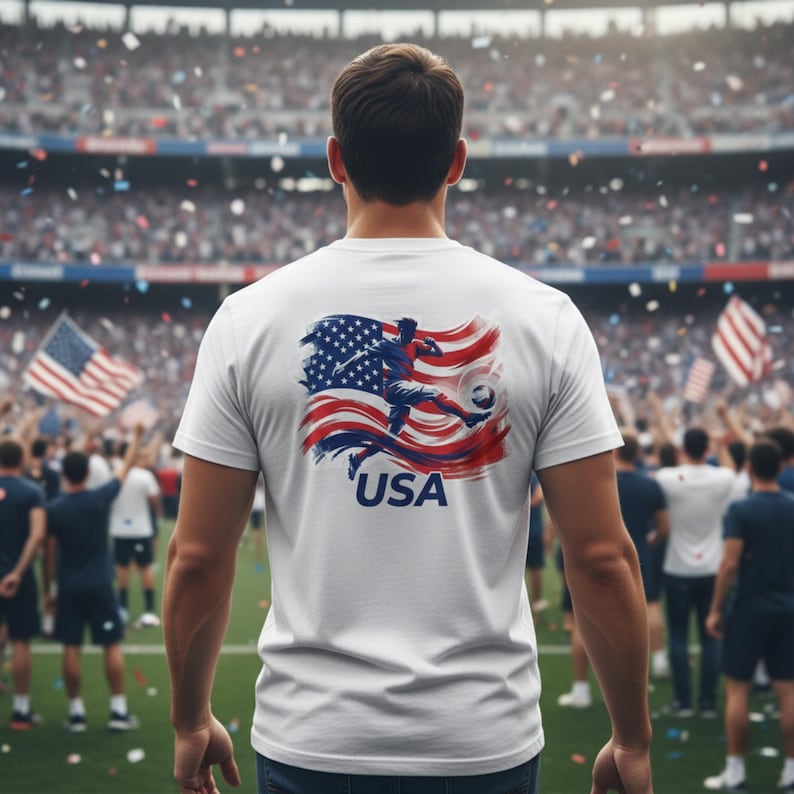 May include: White t-shirt with a graphic of a soccer player kicking a ball, set against a waving American flag design. The word "USA" is printed below the graphic. The image is taken from behind a person in a stadium.
